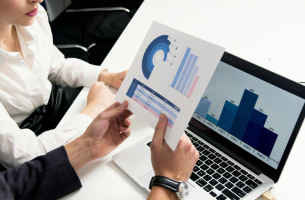 Divi cilvēki skatās grafikus uz papīra lapas un datorā.